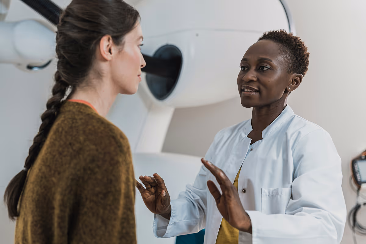 Female doctor in a white coat explaining a medical procedure to a patient in front of medical equipment.