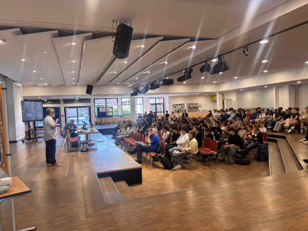 Ein Vortragender spricht in einem großen Saal vor einem aufmerksamen Publikum, das auf Stühlen und Tribünen sitzt.