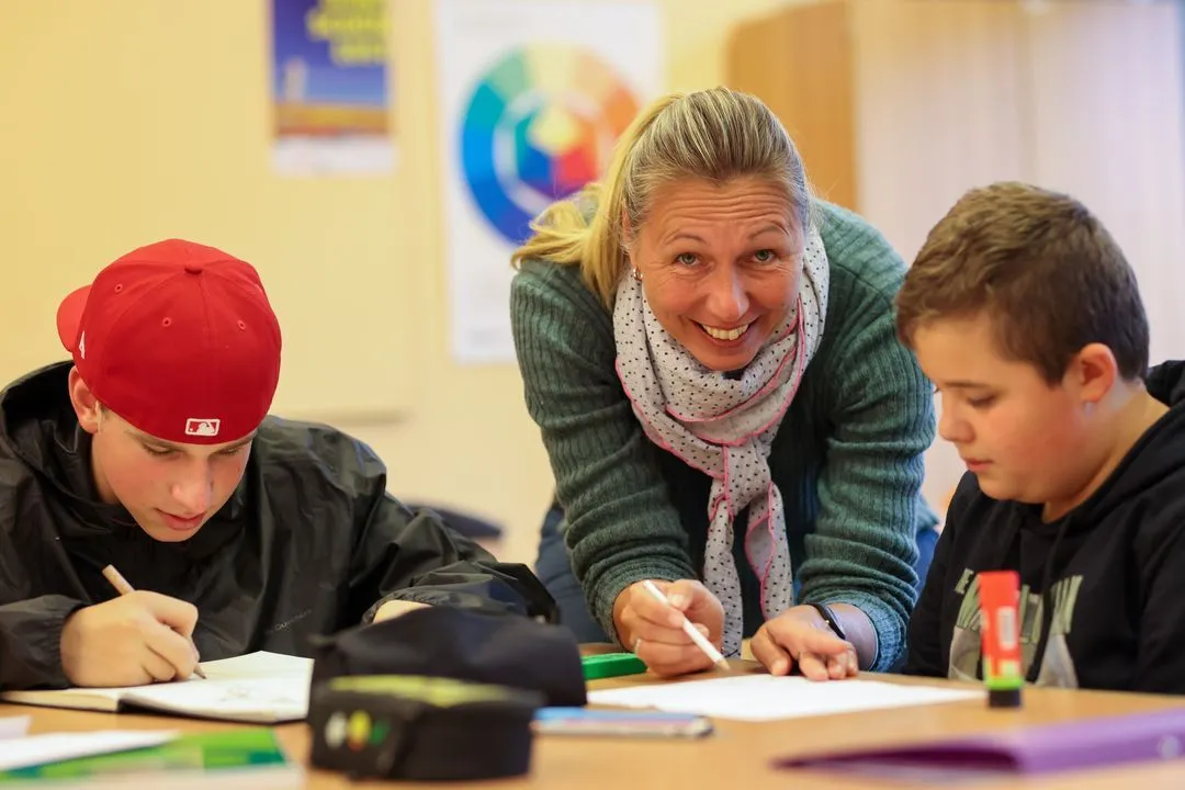 Lehrerin hilft zwei Jungen beim Schreiben an einem Tisch in einem Klassenzimmer.