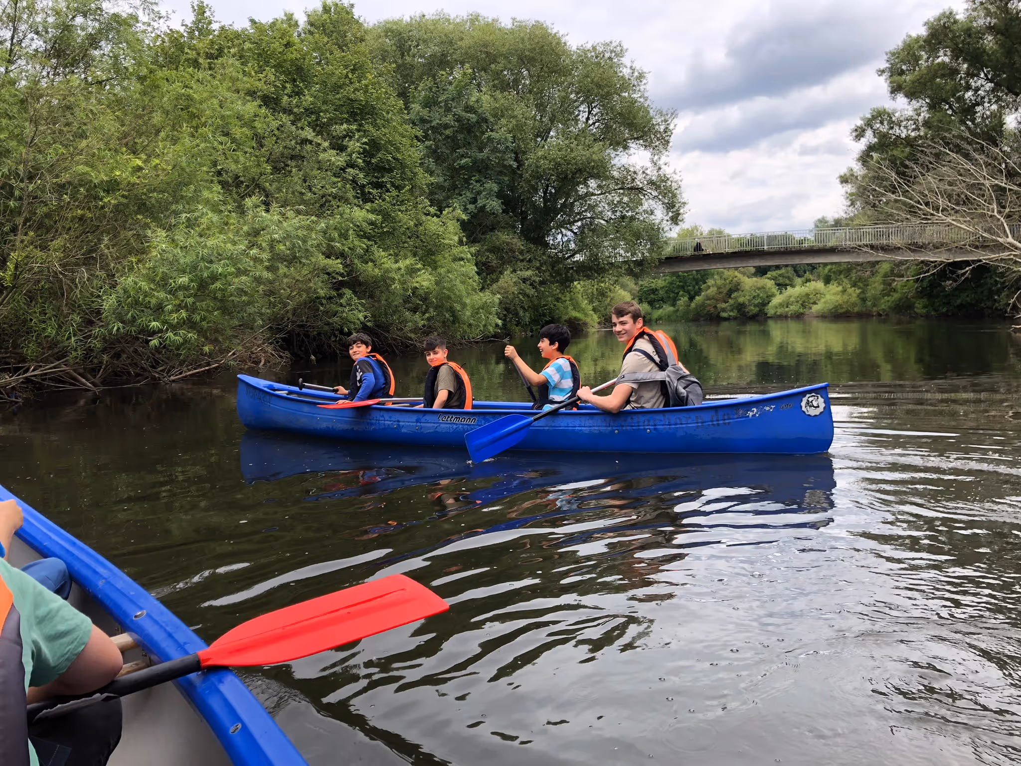 Vier Jugendliche in blauen Kanus paddeln auf einem ruhigen Fluss umgeben von grünen Bäumen und einer Fußgängerbrücke im Hintergrund.