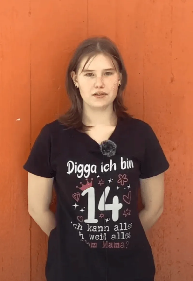 Jugendliche vor orangefarbener Wand trägt ein schwarzes T-Shirt mit dem Text „Digga ich bin 14, ich kann alles, ich weiß alles, warum Mama?“
