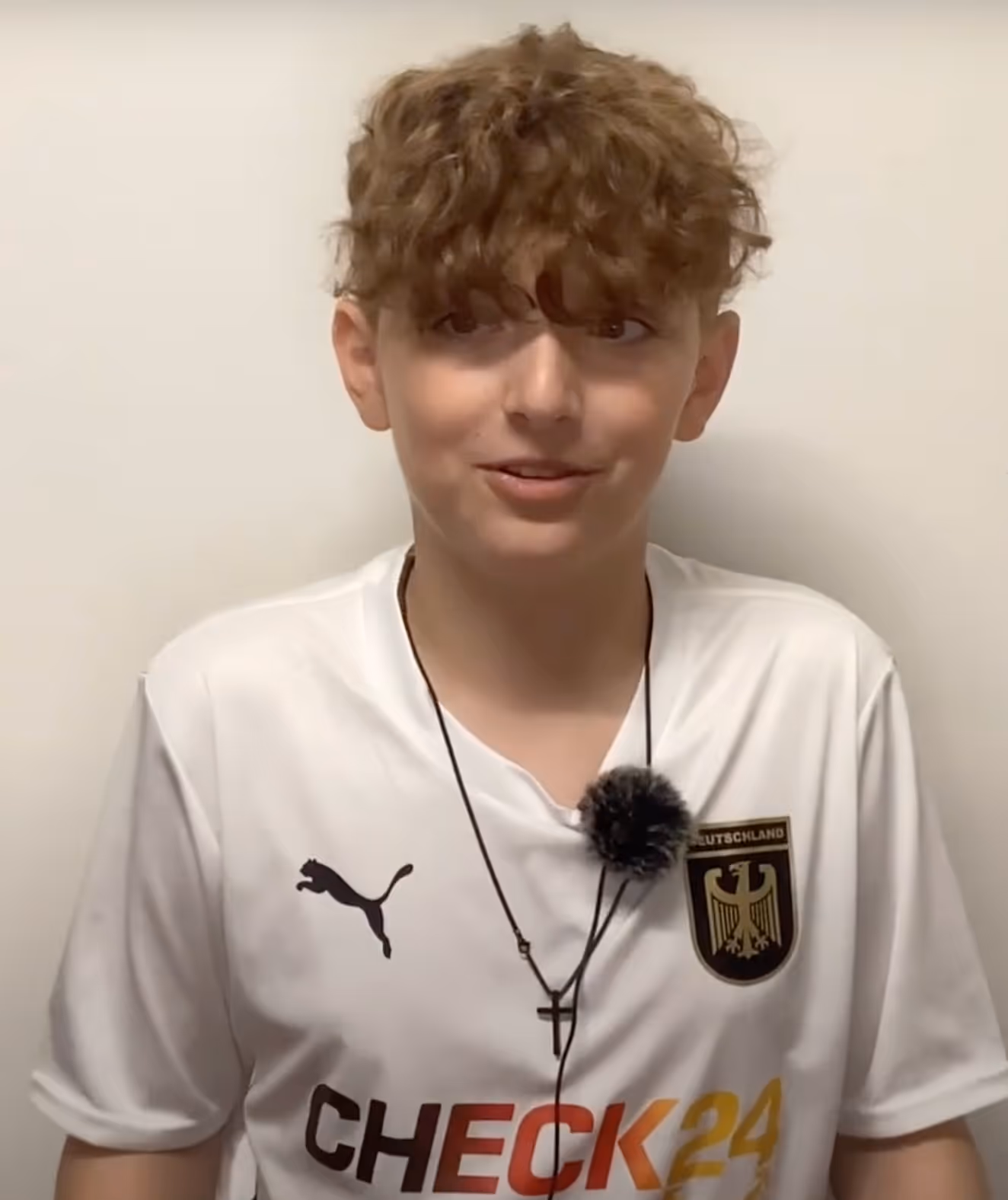 Junge mit lockigem Haar trägt ein weißes Deutschland-Fußballtrikot mit Puma-Logo und Check24-Werbung vor weißem Hintergrund.