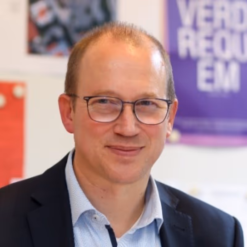 Mann mit Brille, blauem Hemd und dunklem Sakko lächelt leicht in einem Büro mit bunten Postern an der Wand.