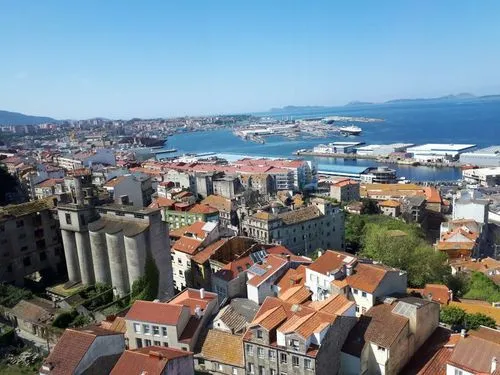 Blick auf eine Hafenstadt mit roten Dächern, Gebäuden, Silos und einem weitläufigen Hafen mit Schiffen unter blauem Himmel.