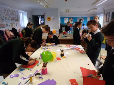 Schüler in Schuluniformen arbeiten gemeinsam an Bastelprojekten mit farbigem Papier und Luftballons in einem Klassenraum.