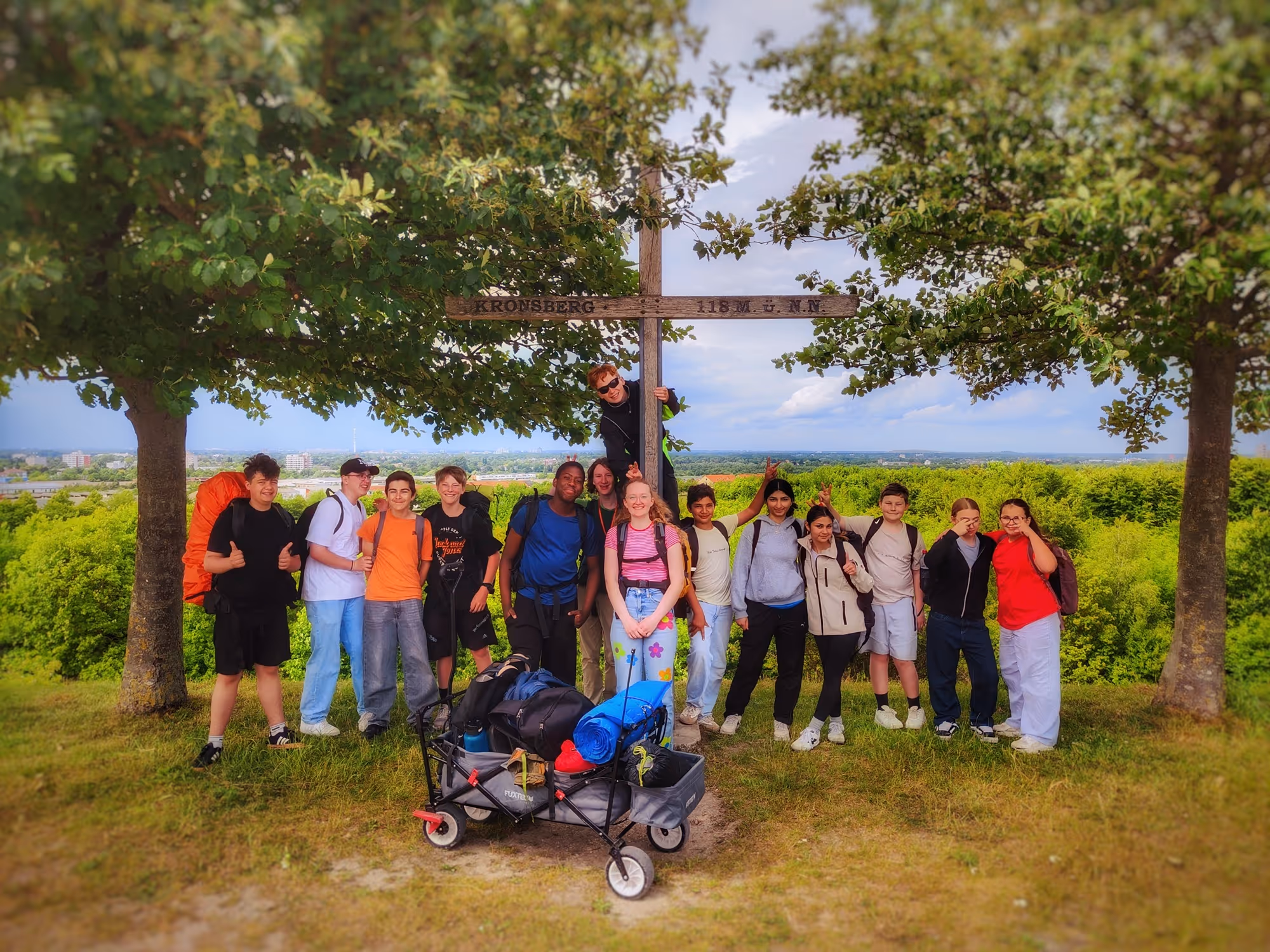 Gruppe von Jugendlichen posiert um ein Holzkreuz auf einem Hügel mit Blick auf eine Stadt und grüne Landschaft.