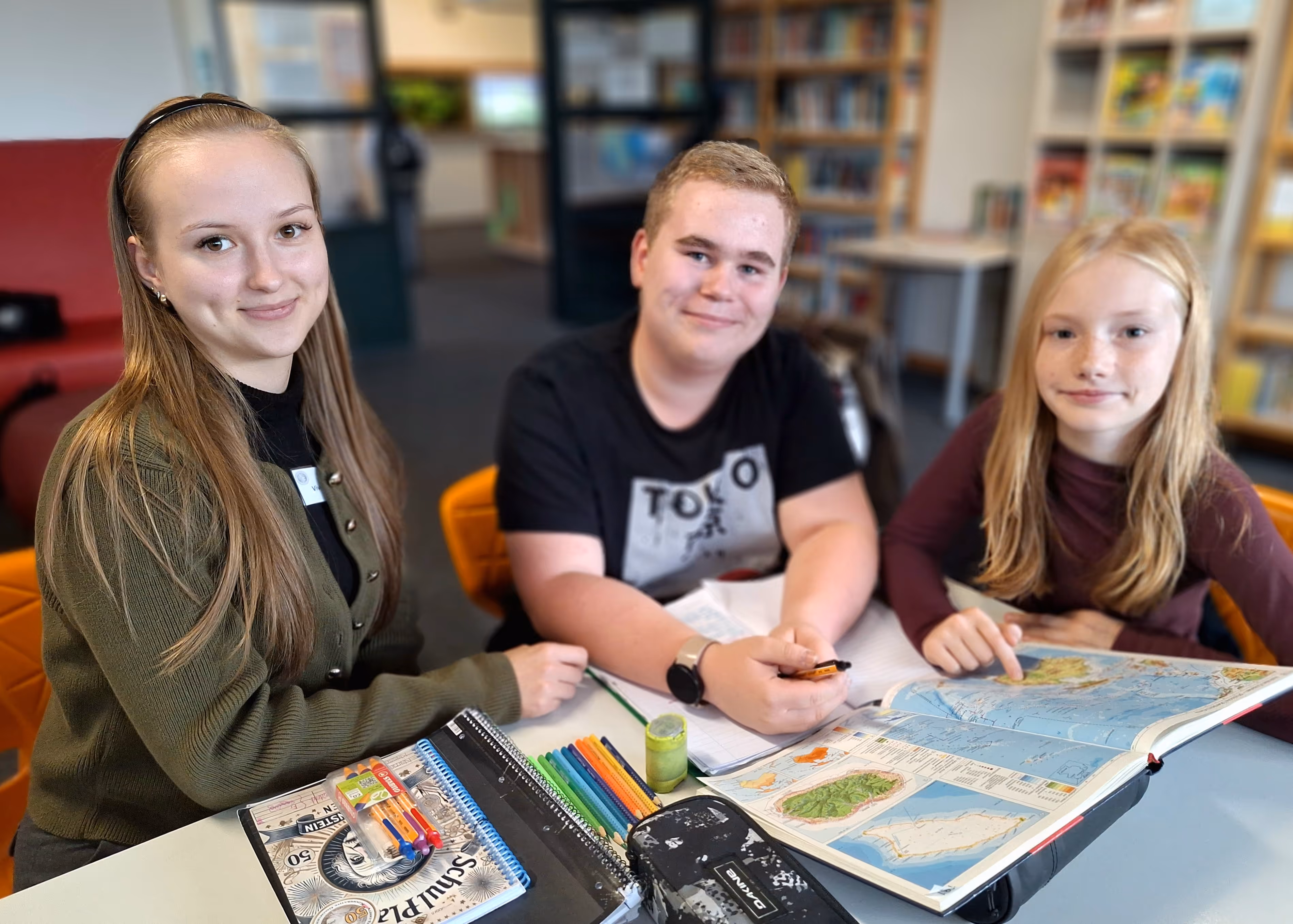 Drei Jugendliche sitzen an einem Tisch in einer Bibliothek und studieren zusammen an einem aufgeschlagenen Atlas.