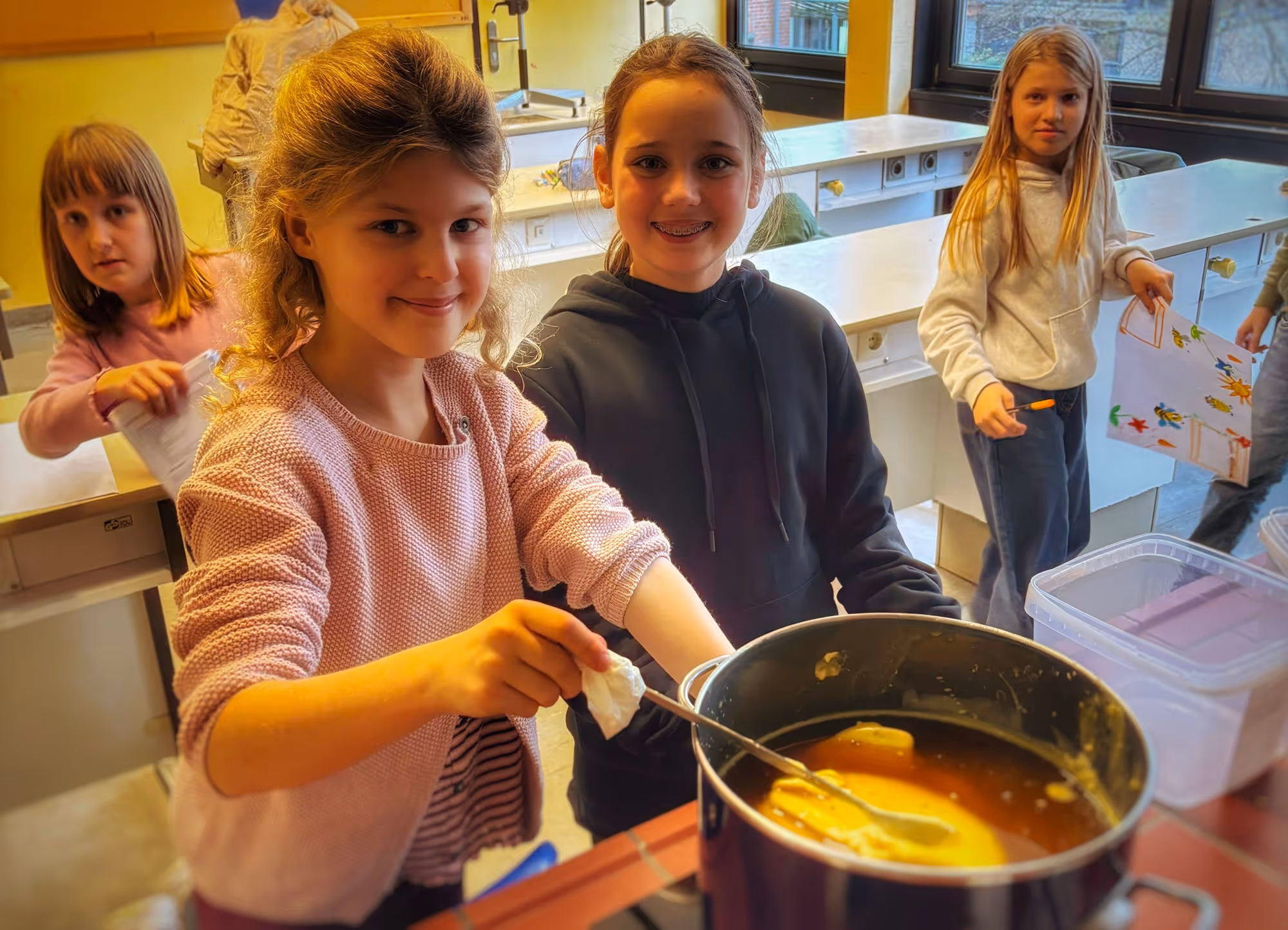 Vier Mädchen in einem Klassenzimmer, zwei davon rühren in einem Topf auf dem Herd, im Hintergrund hält ein Mädchen ein gemaltes Bild.