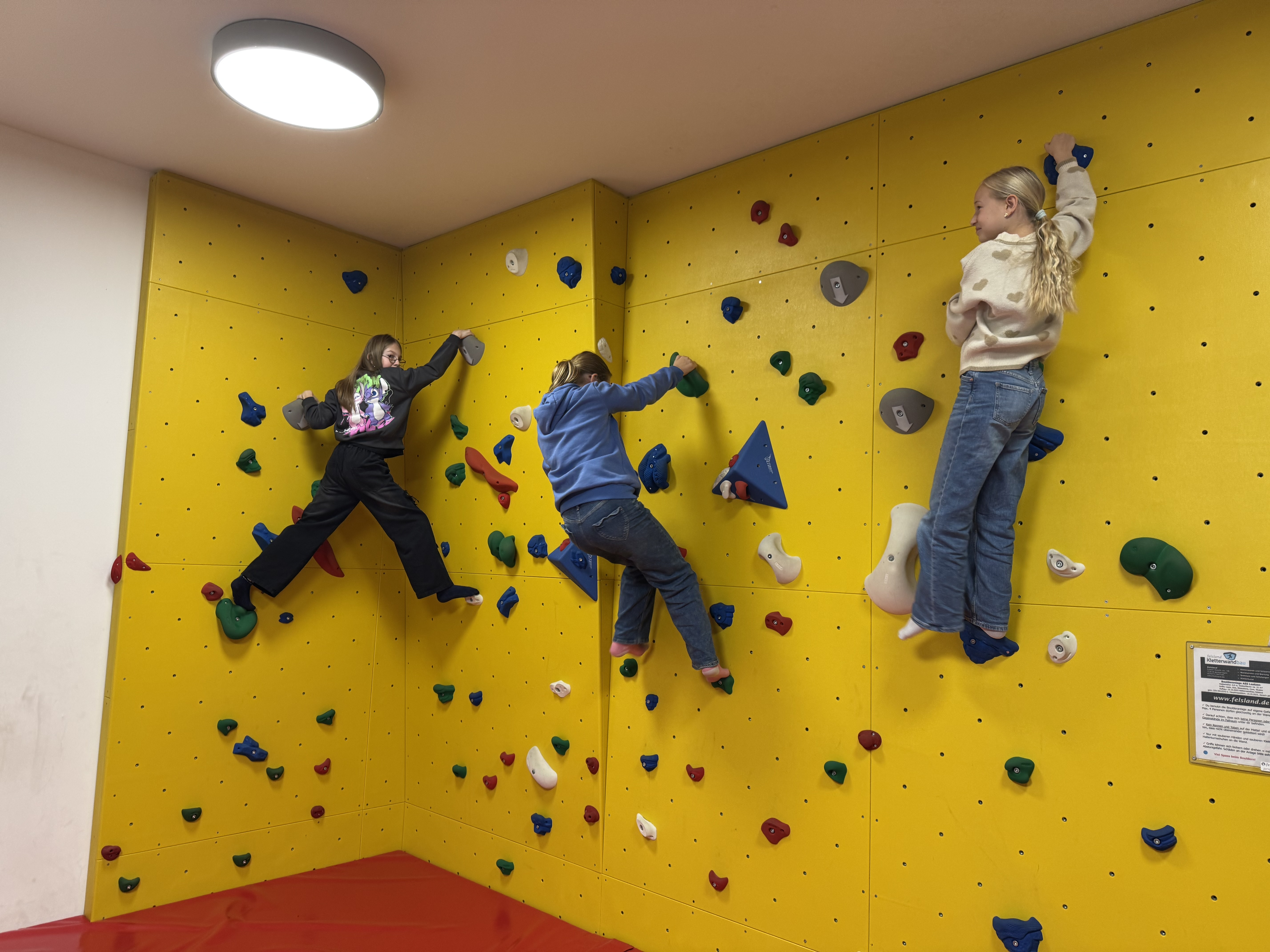 Drei Mädchen klettern an einer gelben Kletterwand mit bunten Griffen in einer Halle.
