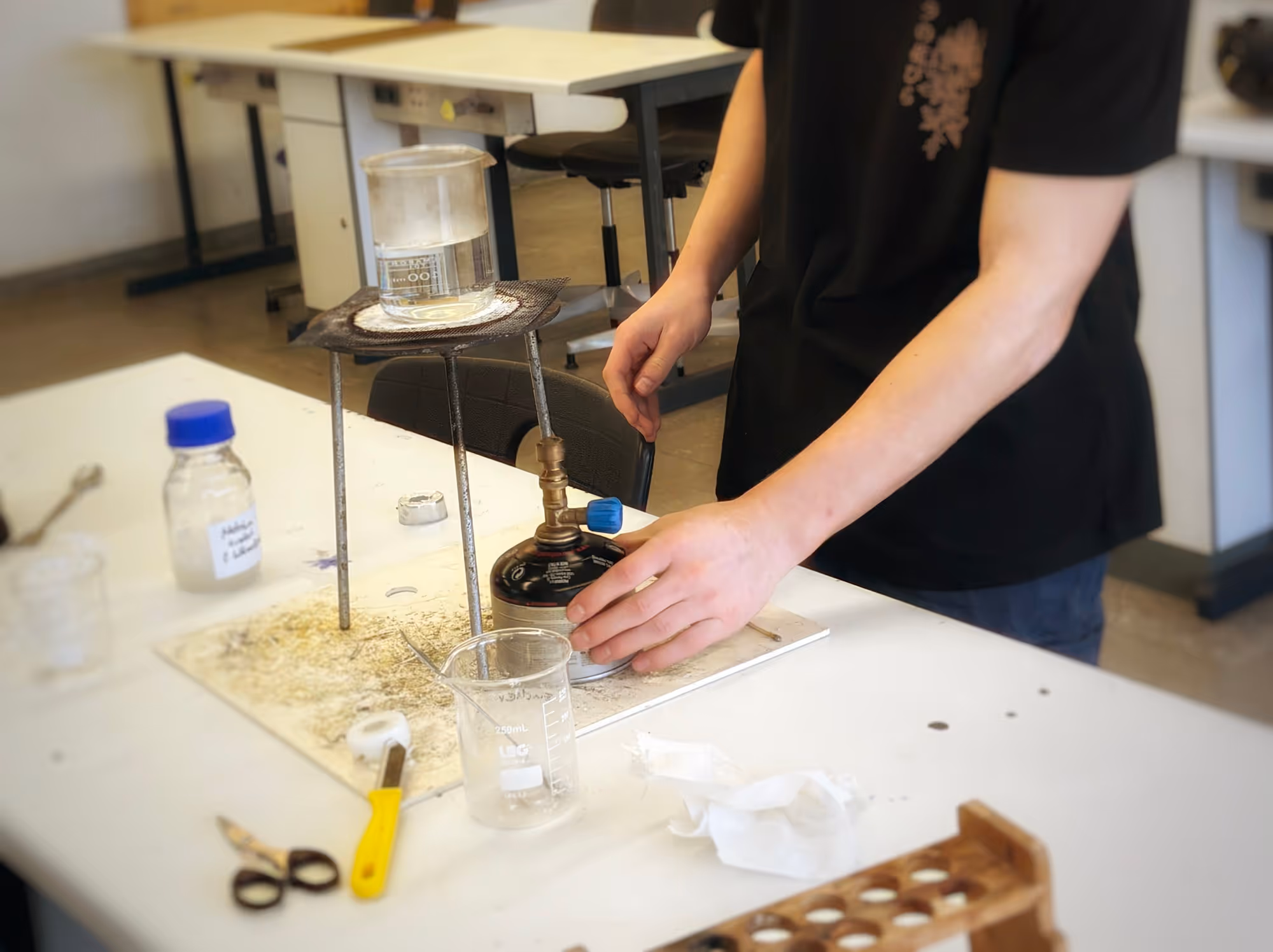 Jemand bedient eine Gasflamme unter einem Bunsenbrenner, der ein mit Wasser gefülltes Becherglas in einem Labor erhitzt.