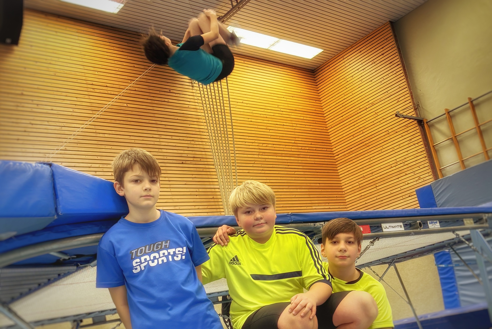 Drei Jungen sitzen an einem Trampolin in einer Turnhalle, während ein vierter Junge eine Rolle in der Luft über dem Trampolin macht.