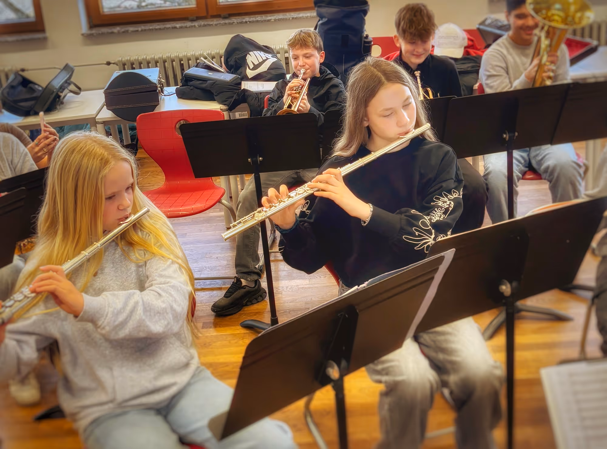 Jugendliche in einem Musikraum spielen Querflöte und Trompete, sitzen auf Stühlen mit Notenständern vor sich.