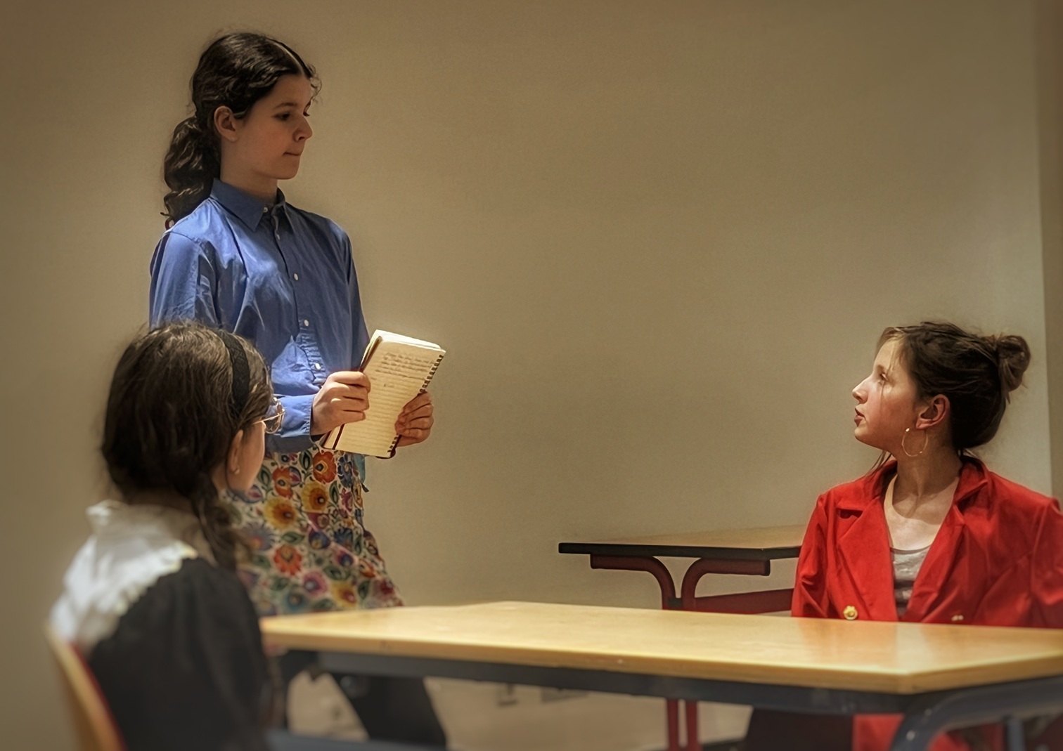 Drei Mädchen in einem Klassenzimmer, eine steht mit einem Notizblock, die anderen sitzen an Tischen und hören zu.