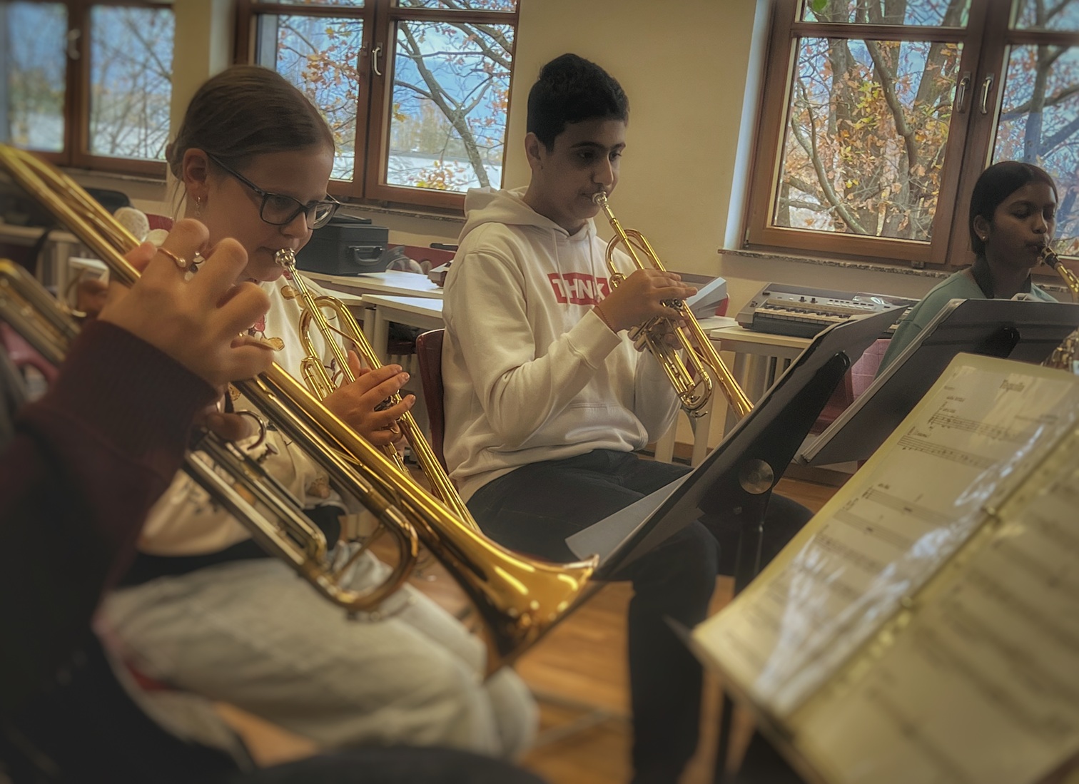 Jugendliche spielen Trompeten in einem Raum mit großen Fenstern und Notenständern.