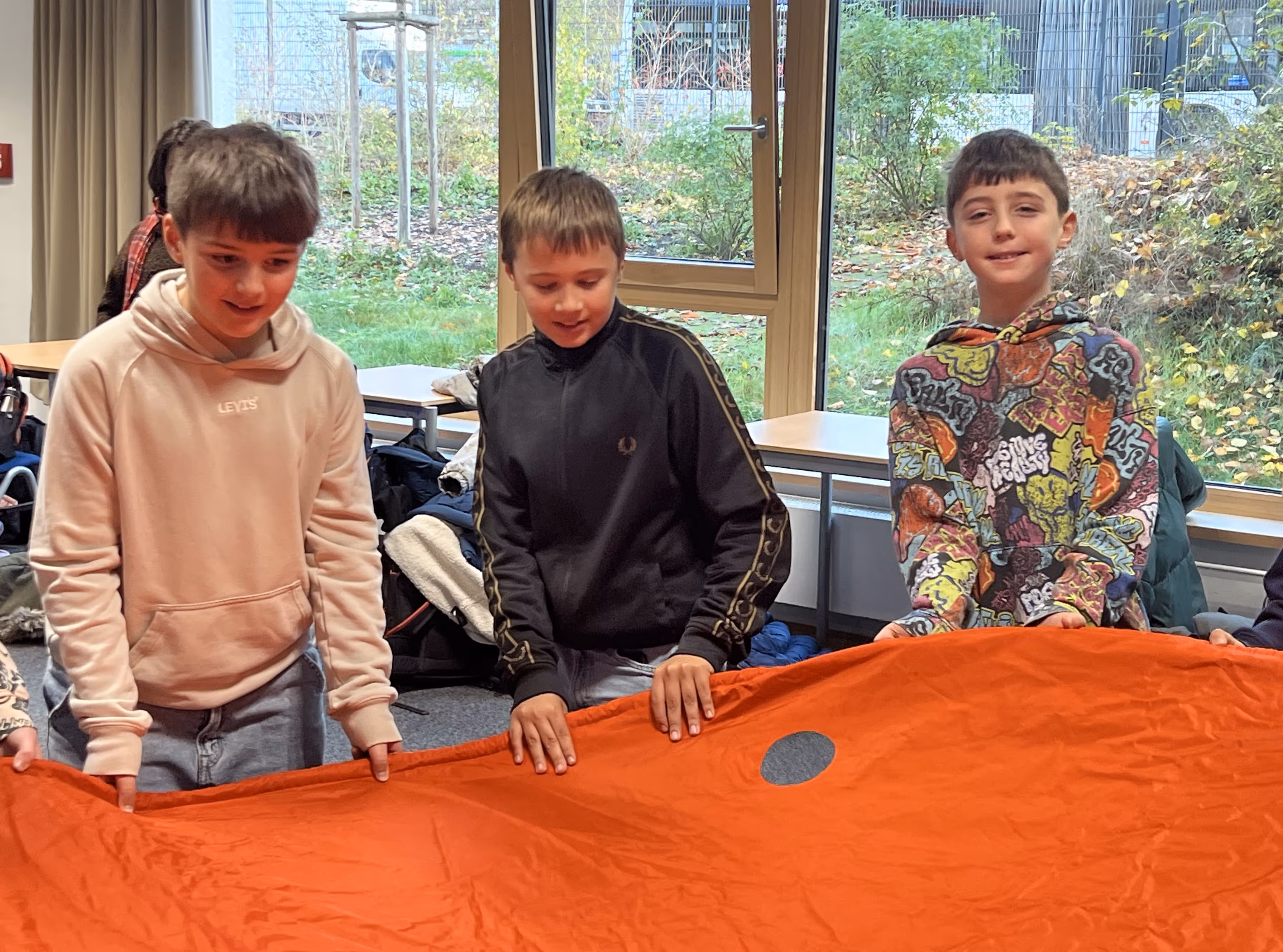 Drei Jungen stehen in einem Klassenraum und halten gemeinsam eine große orangefarbene Decke.