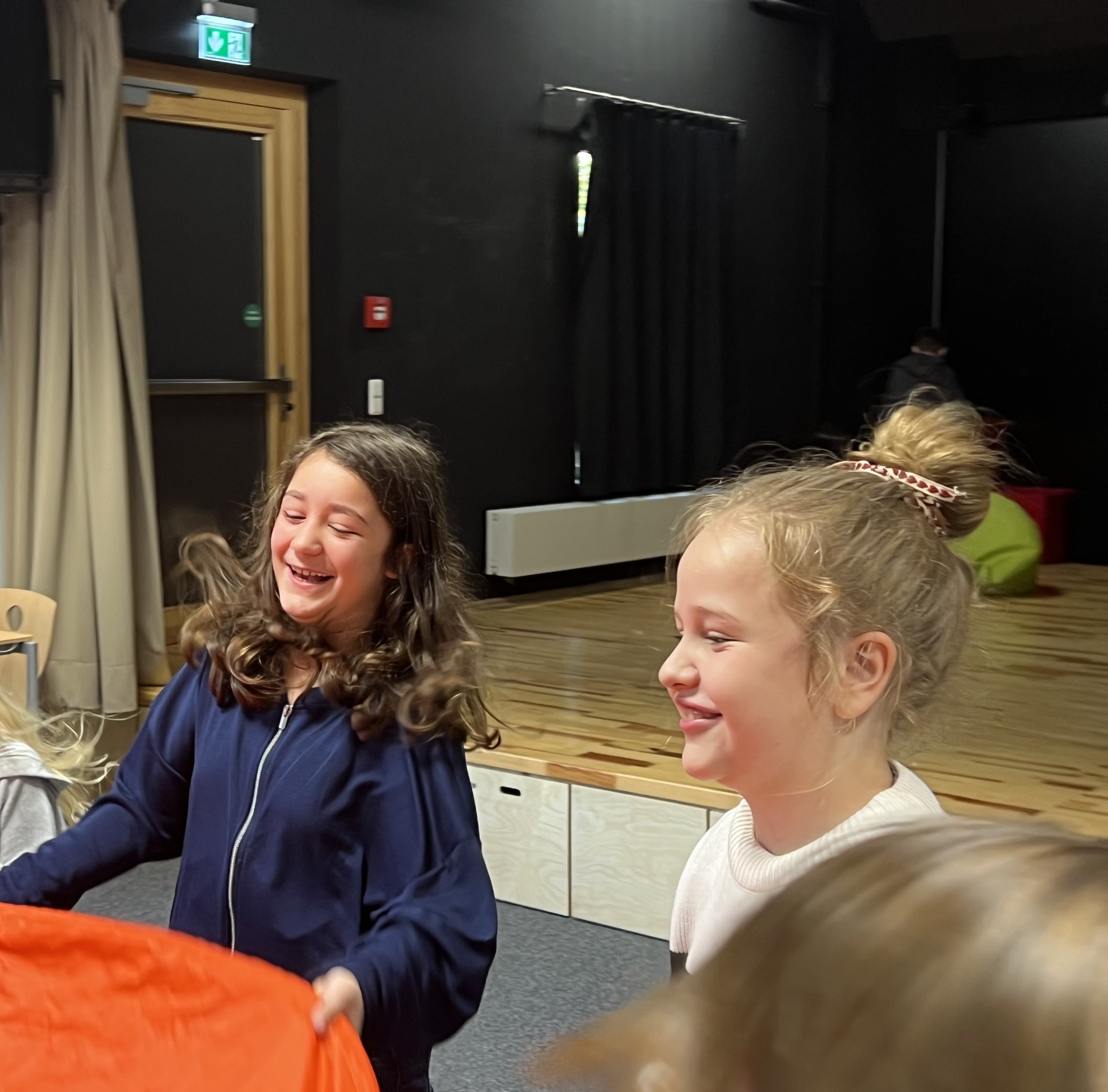 Zwei lachende Mädchen spielen zusammen in einem Innenraum mit Holzfußboden und schwarzer Wand.