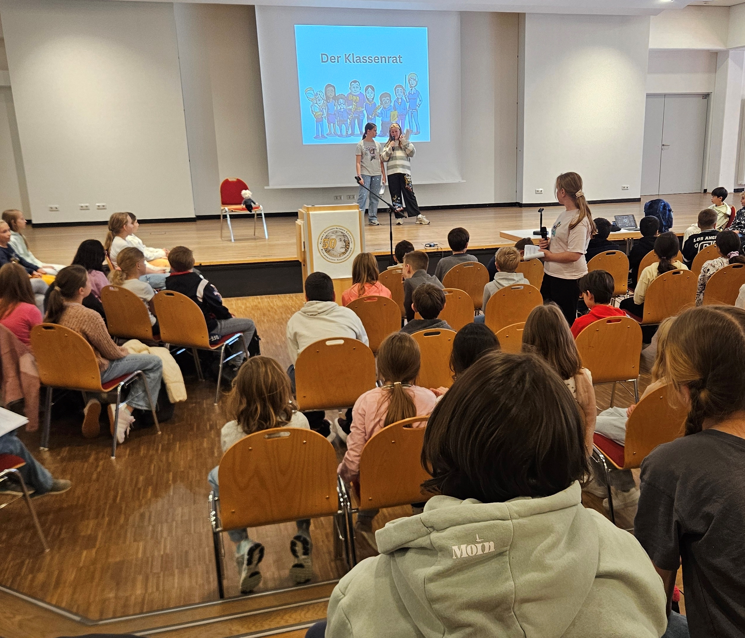 Schüler, die in einem Klassenraum an einem Vortrag zum Thema Klassenrat teilnehmen, während zwei Kinder auf der Bühne sprechen.