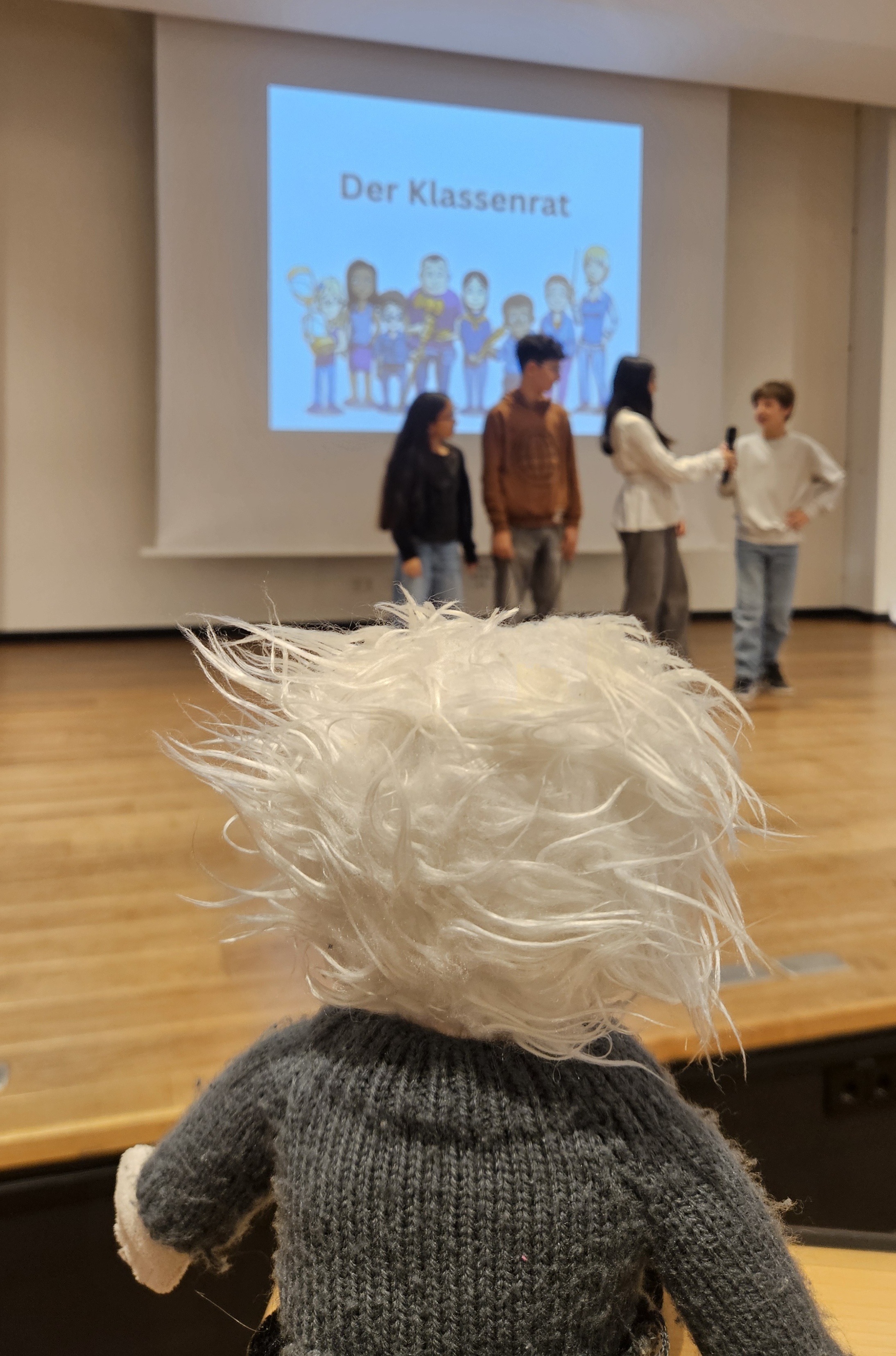 Ein Kuscheltier mit weißem zerzaustem Haar schaut auf eine Bühne, auf der vier Kinder vor einer Leinwand mit der Aufschrift ‚Der Klassenrat‘ stehen.