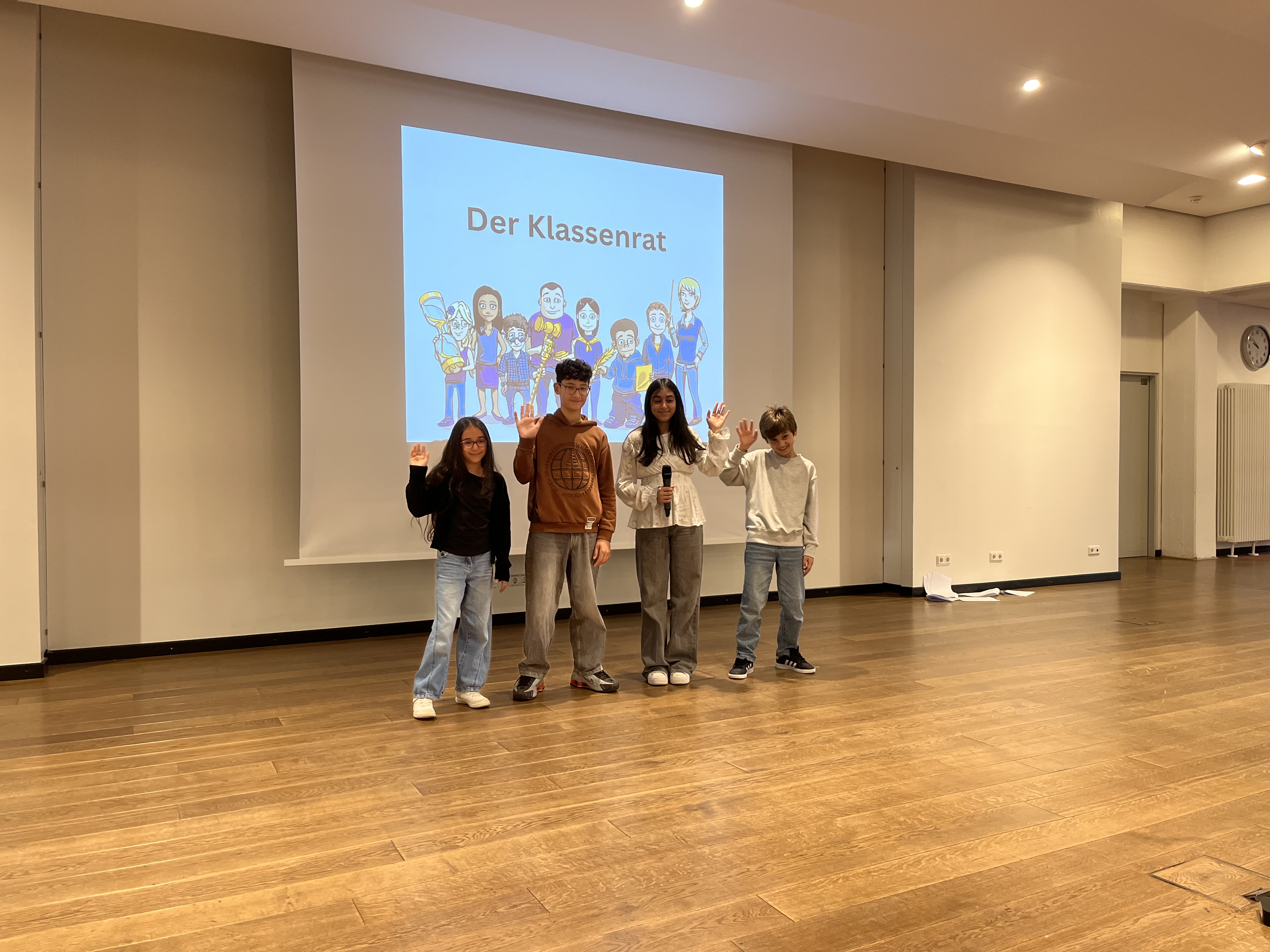 Vier Kinder stehen vor einer Leinwand mit der Aufschrift "Der Klassenrat" und heben die Hand.