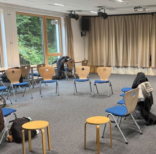 Stuhlkreis mit blauen Stühlen und Holzhockern in einem Raum mit großen Fenstern und beige Vorhängen.