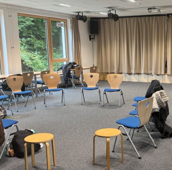 Stuhlkreis mit blauen Stühlen und Holzhockern in einem Raum mit großen Fenstern und beige Vorhängen.