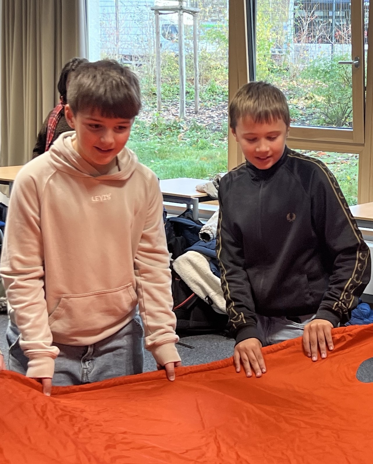 Zwei Jungen halten eine große orangefarbene Decke, im Hintergrund Fenster mit Gartenblick.