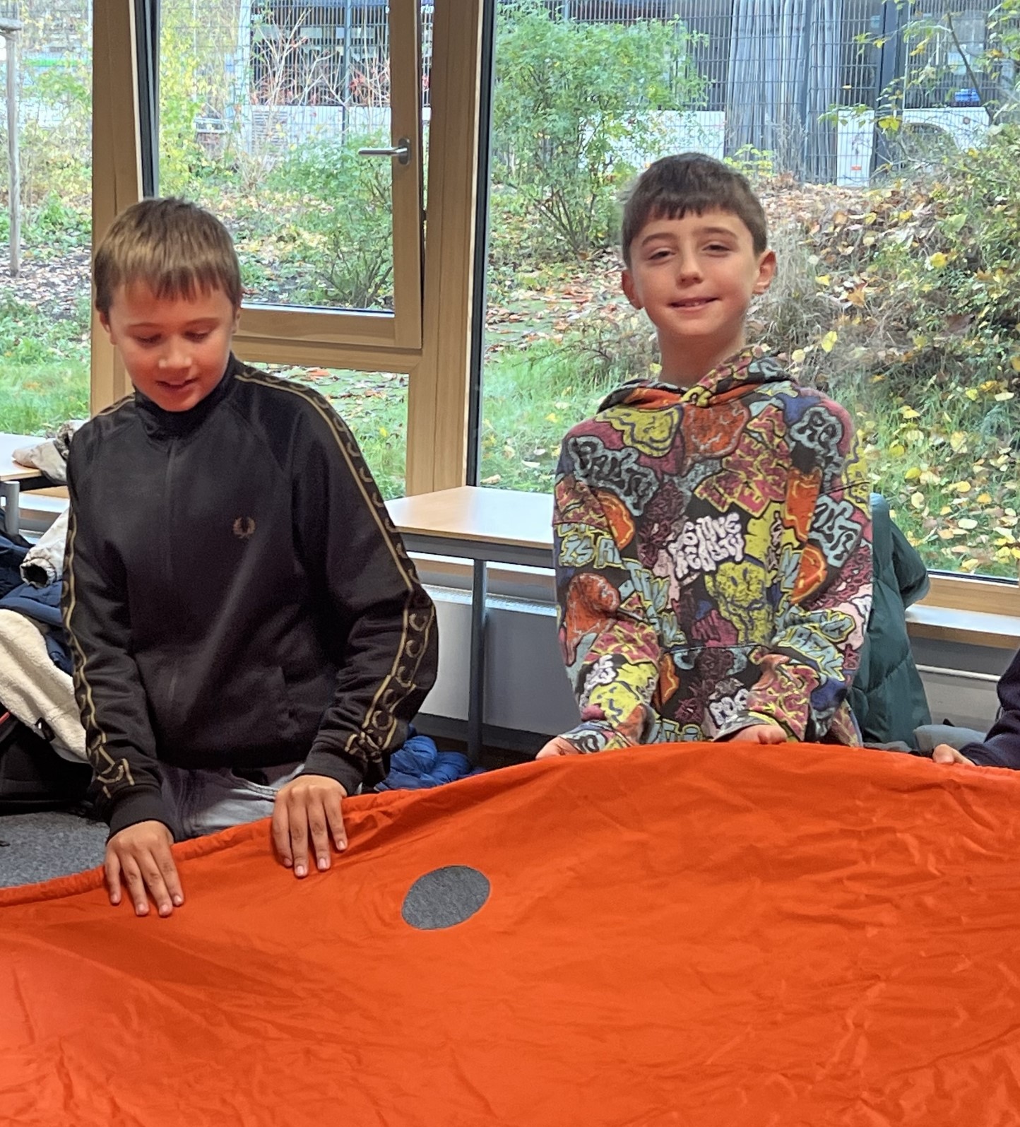 Zwei Jungen sitzen vor einem Fenster und halten gemeinsam ein orangefarbenes Tuch mit einem Loch.