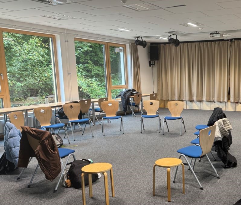 Ein Konferenzraum mit Stühlen und Hockern in einem Kreis, einige Stühle haben Jacken über den Rückenlehnen.