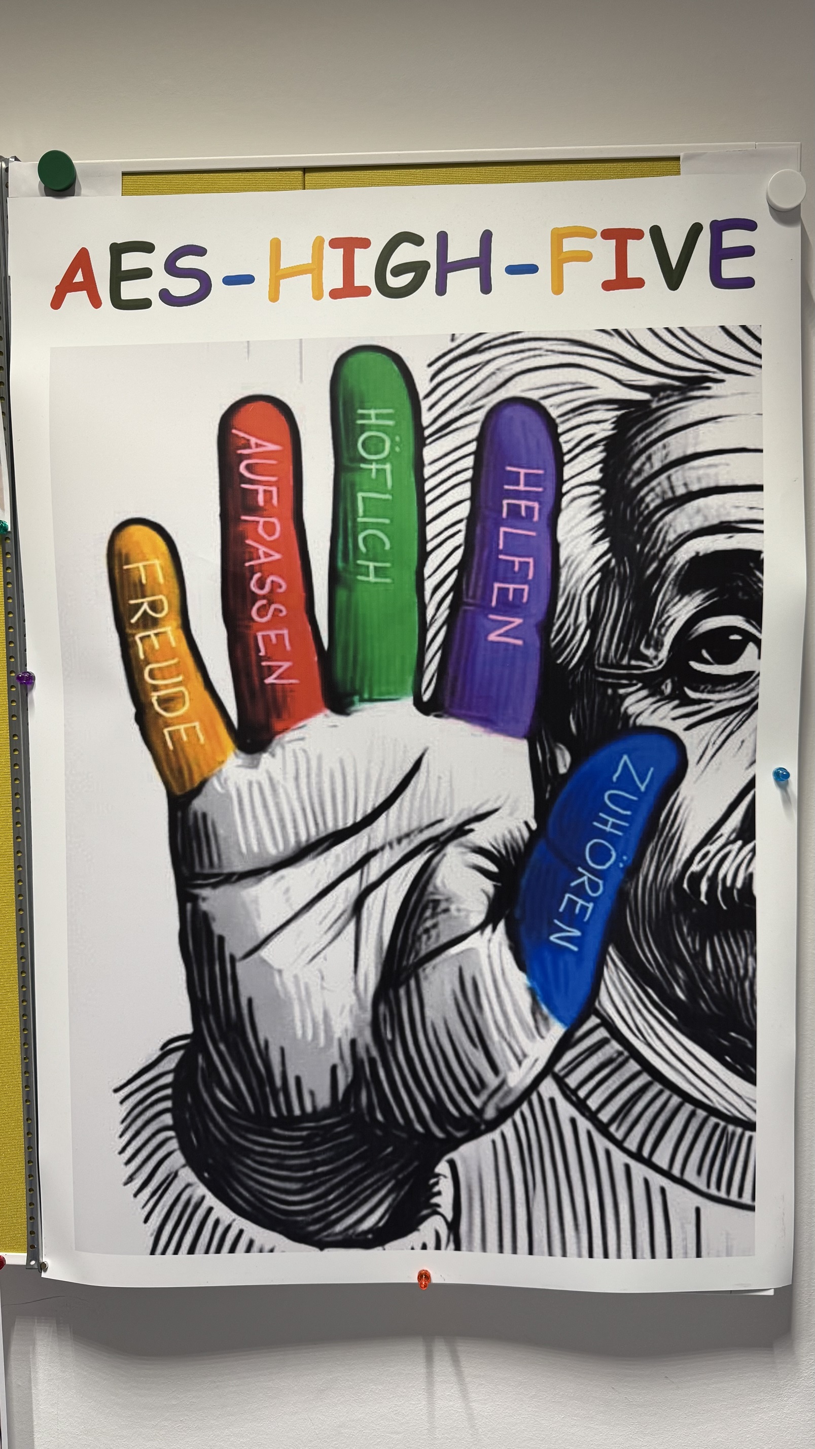Poster mit der Aufschrift AES-HIGH-FIVE und einer gezeichneten Hand, deren Finger bunt eingefärbt und mit Wörtern wie Freude, Aufpassen, Höflich, Helfen und Zuhören beschriftet sind.
