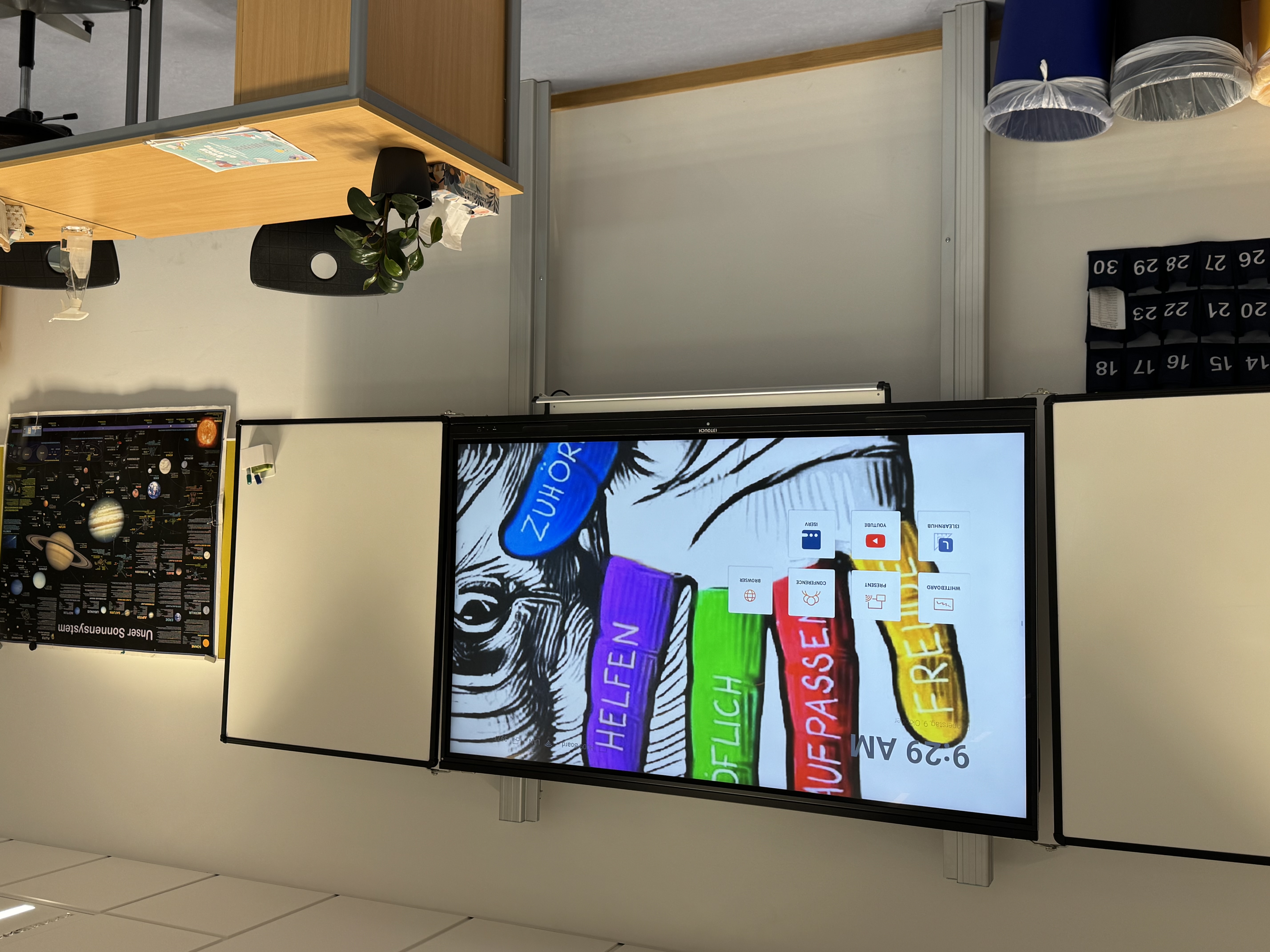 Klassenzimmer mit einem großen interaktiven Bildschirm, der eine farbige Handzeichnung mit deutschen Worten auf den Fingern zeigt, und einem Tisch mit Pflanzen und Reinigungsmitteln.