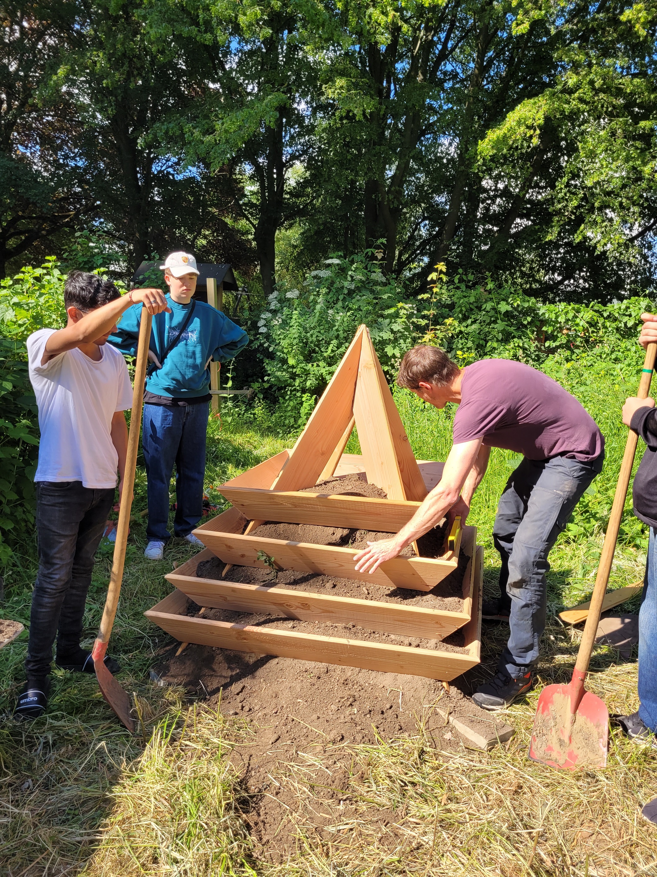 Mehrere Personen bauen eine mehrschichtige hölzerne Pflanzstruktur mit Erde in einem Garten auf.