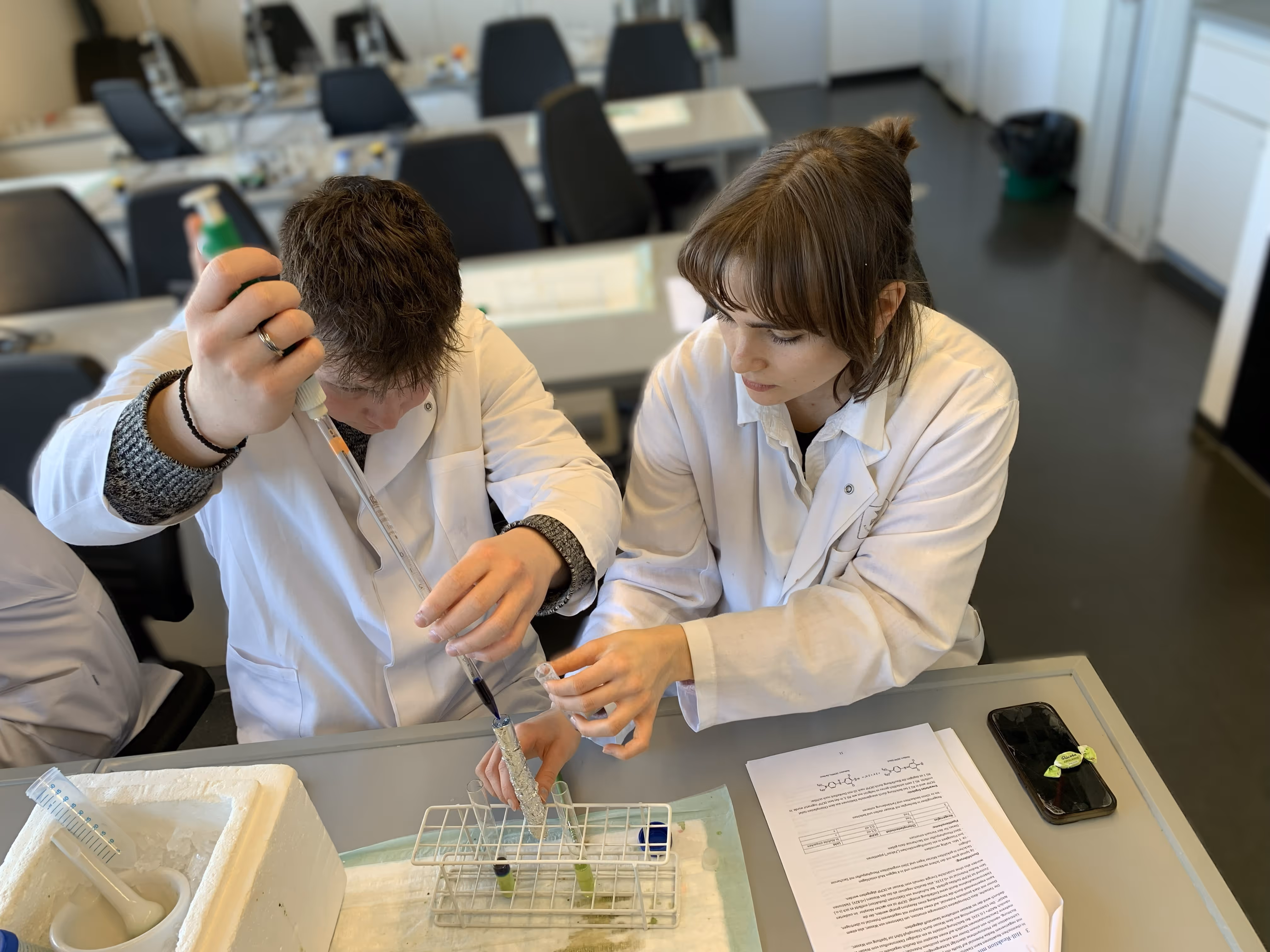 Zwei Schüler tragen weiße Kittel und sitzen an einem Tisch und experimentieren