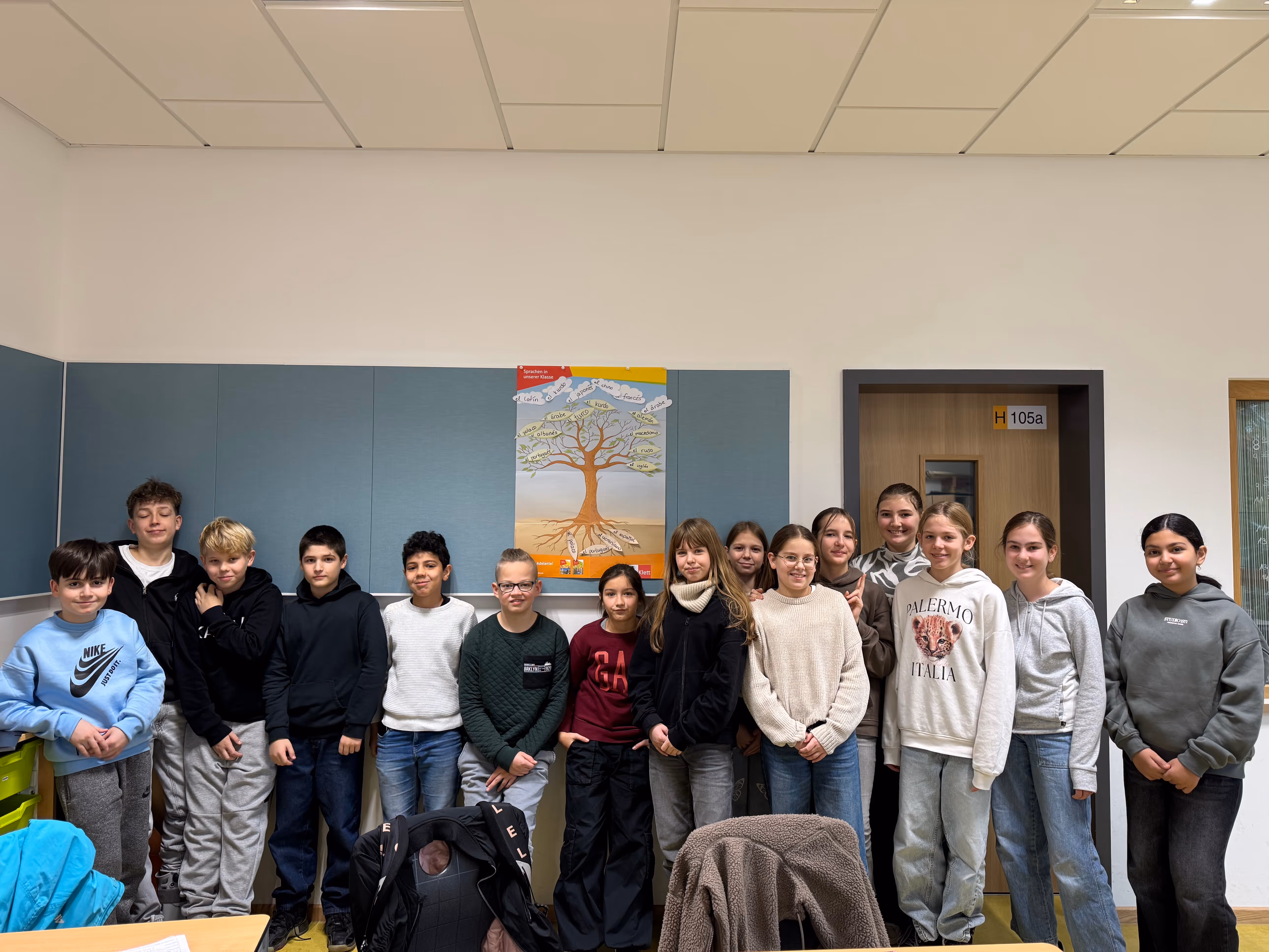 Die Schüler:innen eines Spanischkurses posieren vor einem Poster von einem Sprachenbaum, auf dem alle Sprachen auf Spanisch festgehalten wurden, die in der Klasse gesprochen werden. 