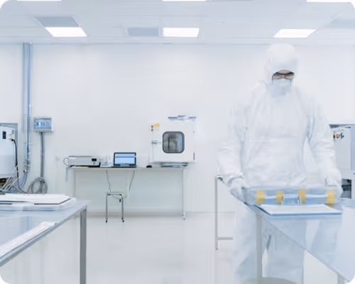 Personne en combinaison de protection blanche dans un laboratoire propre manipulant un équipement sur une table en acier inoxydable.