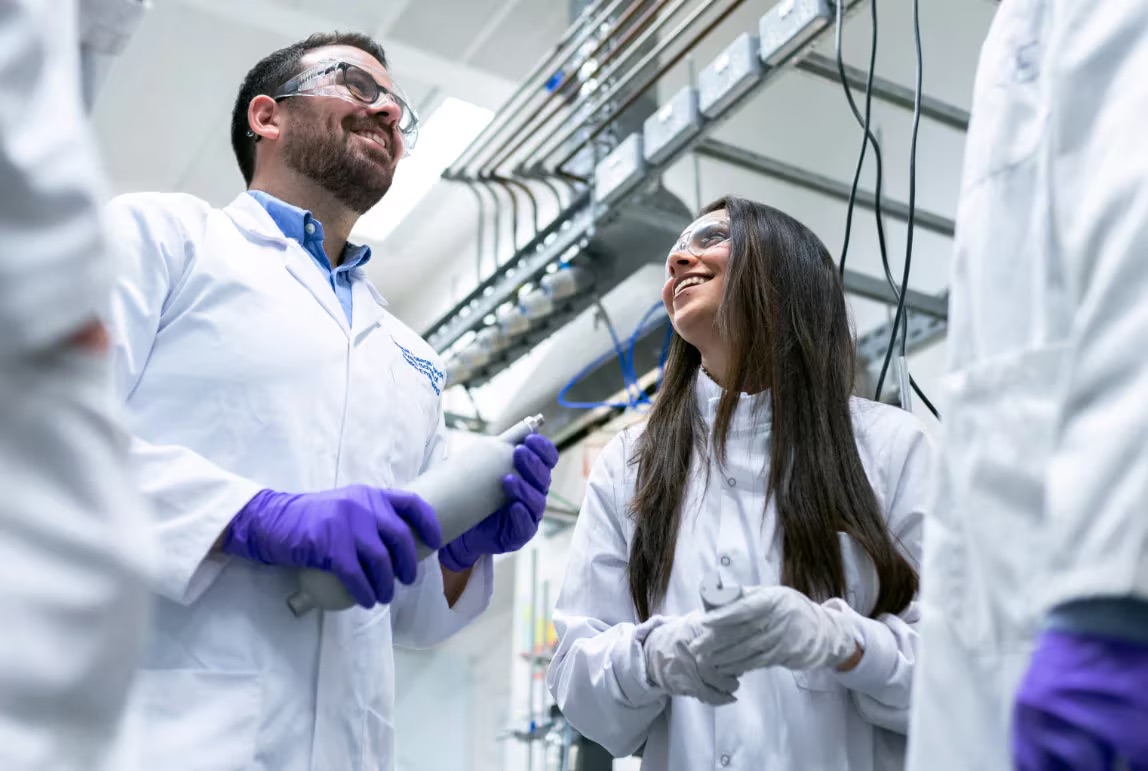 Deux scientifiques en blouse blanche et lunettes de protection discutent dans un laboratoire industriel avec des équipements en arrière-plan.