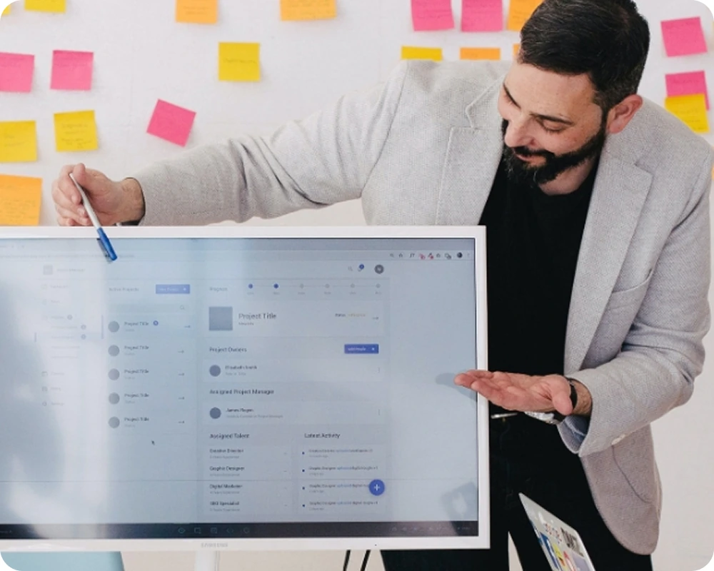 Homme en veste grise présentant un écran d'ordinateur avec un tableau de gestion de projet, mur couvert de notes autocollantes colorées en arrière-plan.