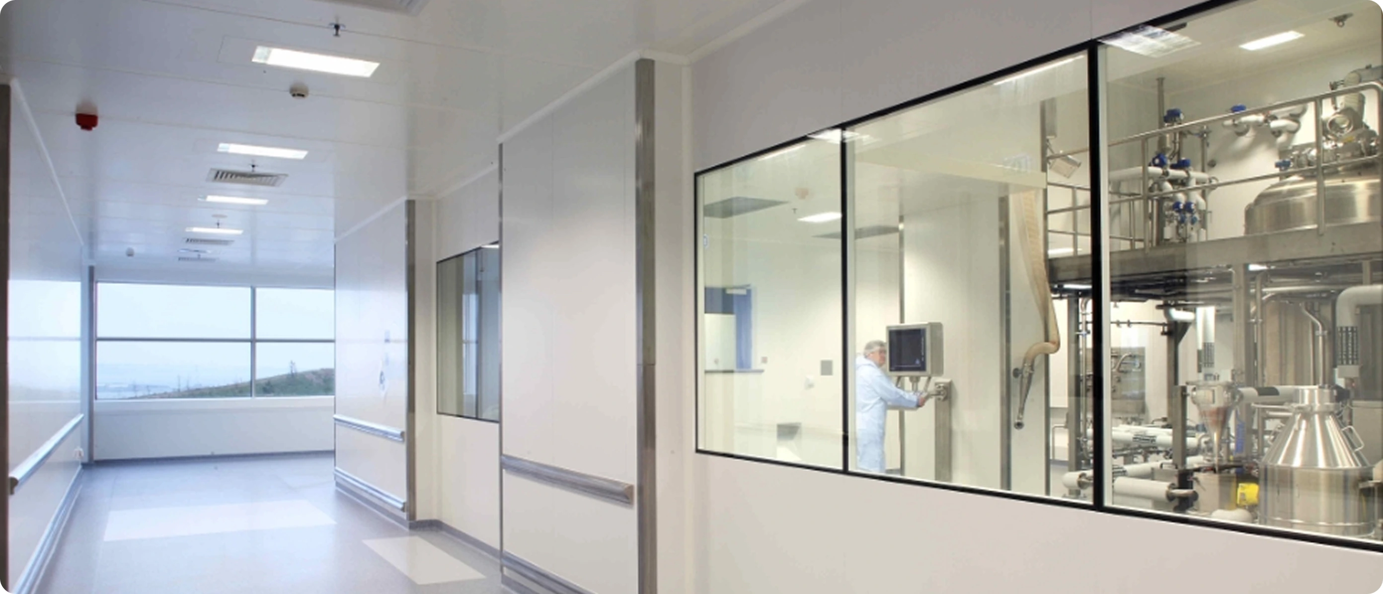 Couloir propre et lumineux avec fenêtres donnant sur une salle de fabrication industrielle où une personne en tenue blanche travaille sur un écran.