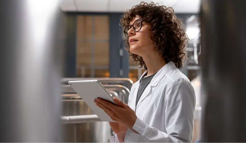 Femme en blouse blanche tenant une tablette dans une usine ou un laboratoire industriel.