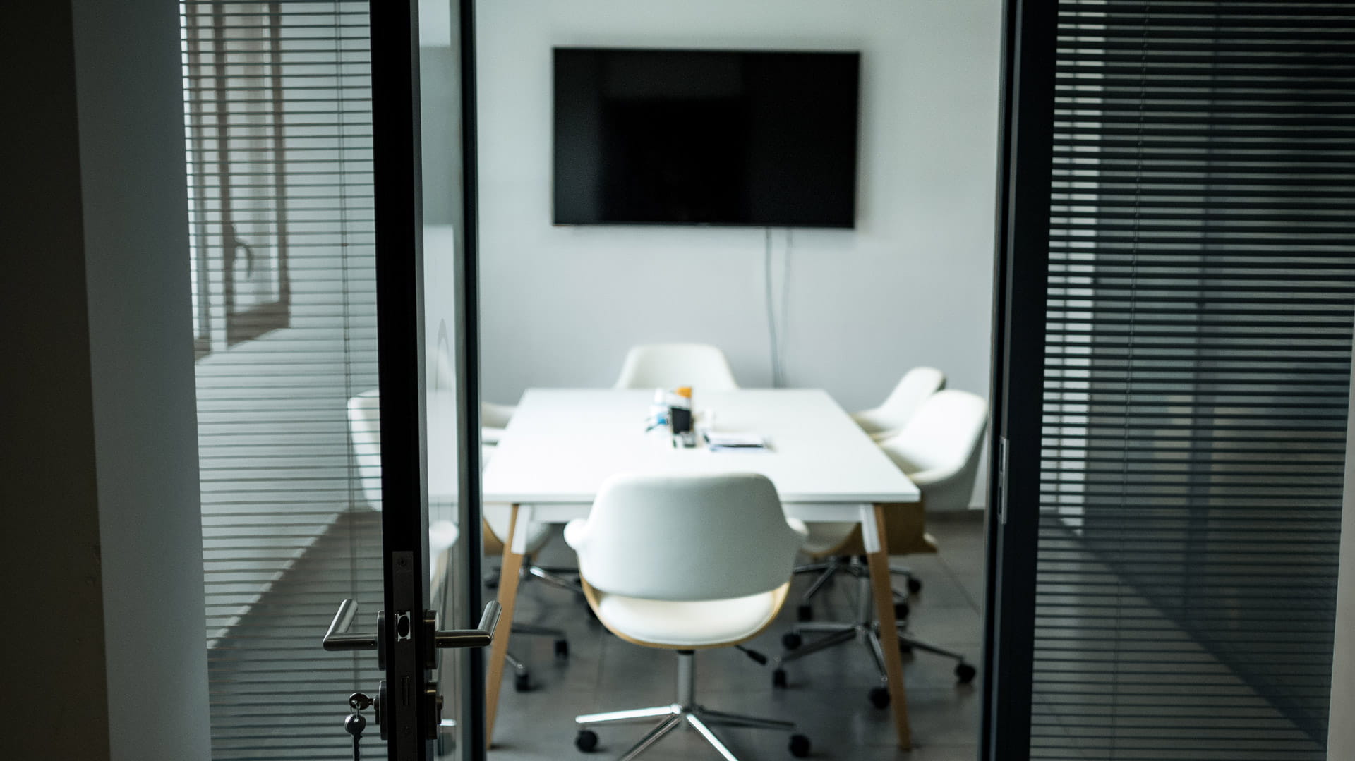 Salle de réunion moderne avec une table blanche entourée de chaises blanches sur roulettes et un écran plat fixé au mur.