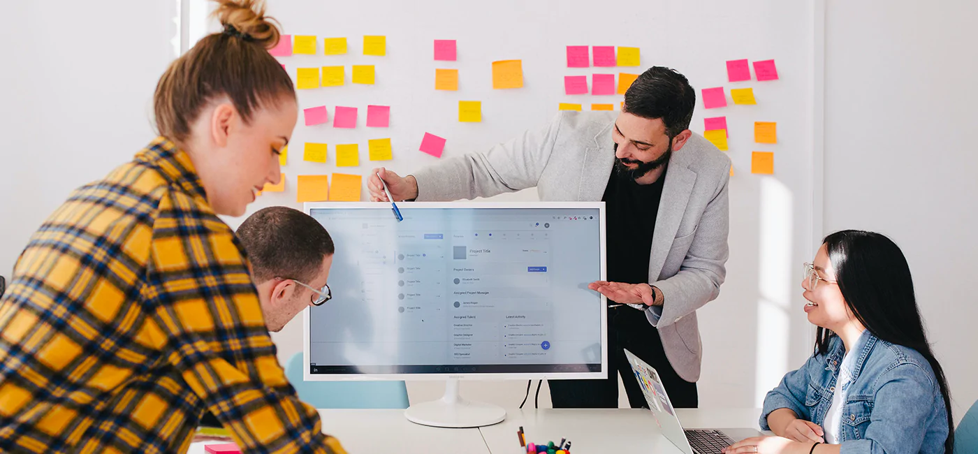 Groupe de quatre personnes en réunion de travail, un homme présentant des informations sur un écran d'ordinateur, tableau blanc avec post-it en arrière-plan.