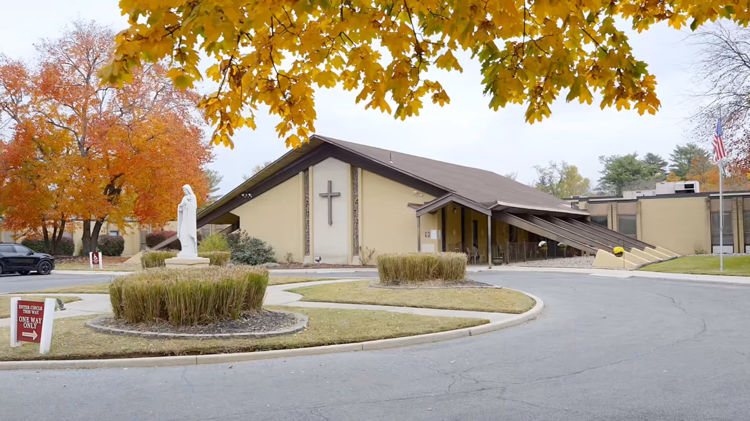 Church building with a cross on the front wall, a statue in the circular driveway, autumn trees with orange and yellow leaves, and an American flag on a flagpole.