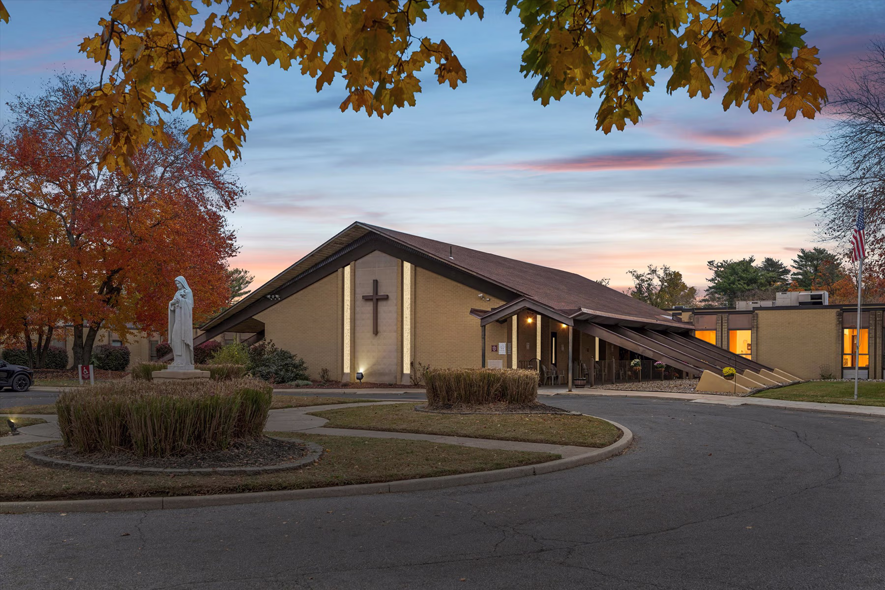 Building with a large cross on the front, a statue of a religious figure in the foreground, autumn trees, and an American flag at sunset.