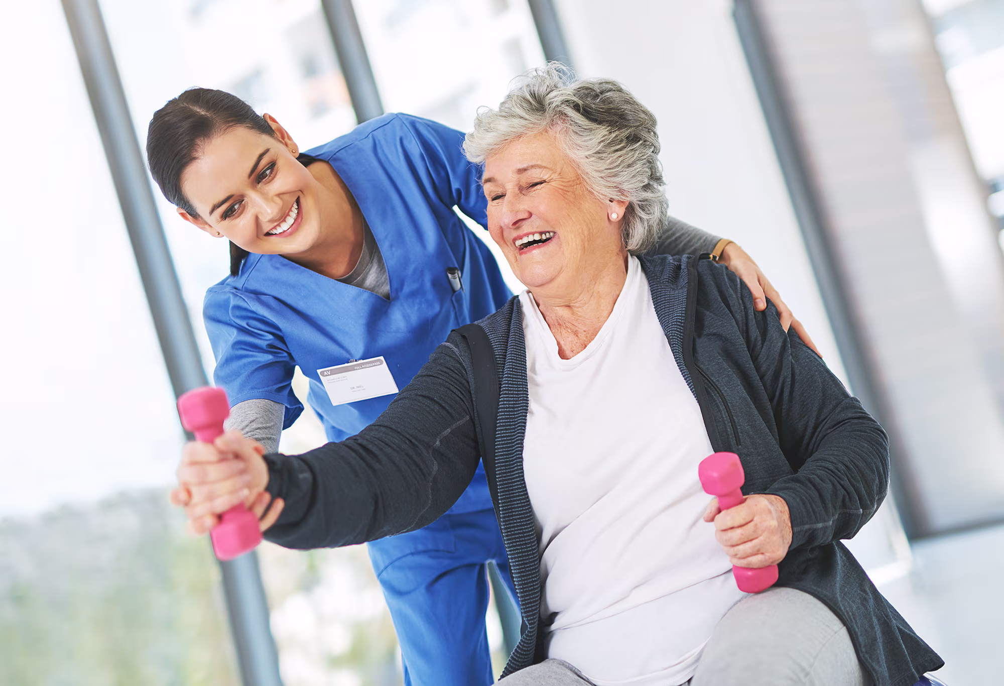 Smiling female nurse in blue scrubs assists elderly woman lifting pink dumbbells during exercise.