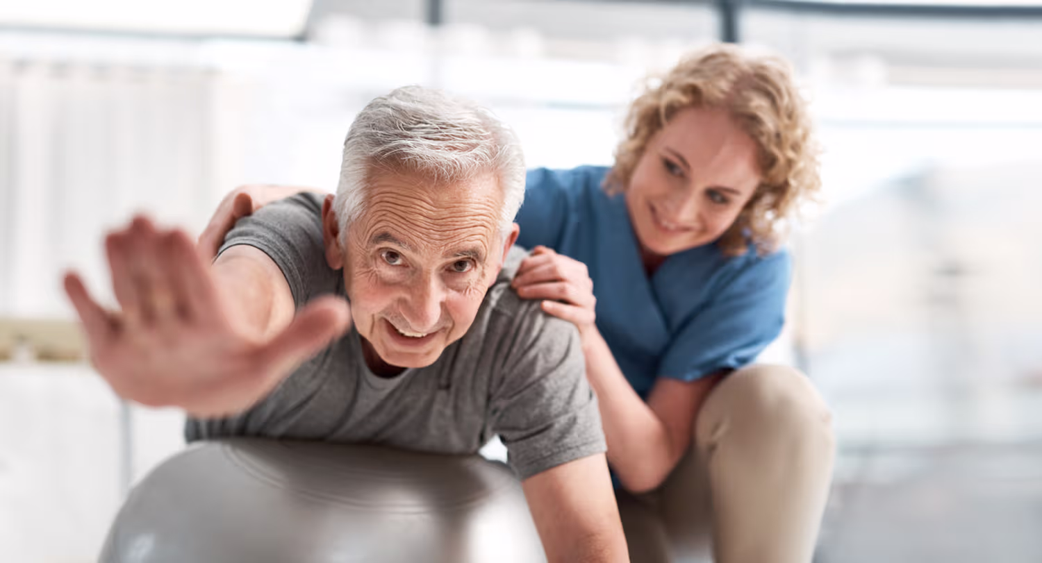 Smiling elderly man stretching on exercise ball with support from a female physical therapist in a bright room.