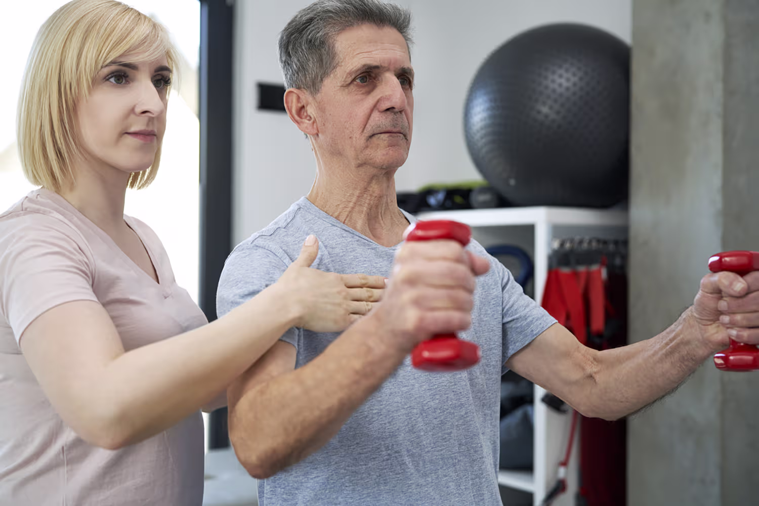Woman assisting an older man lifting red dumbbells during a workout session.