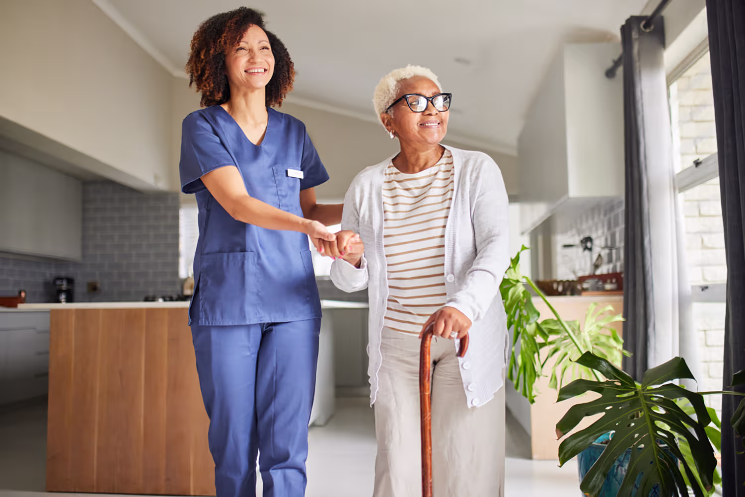 Smiling nurse in blue scrubs assisting elderly woman with a cane walking indoors.