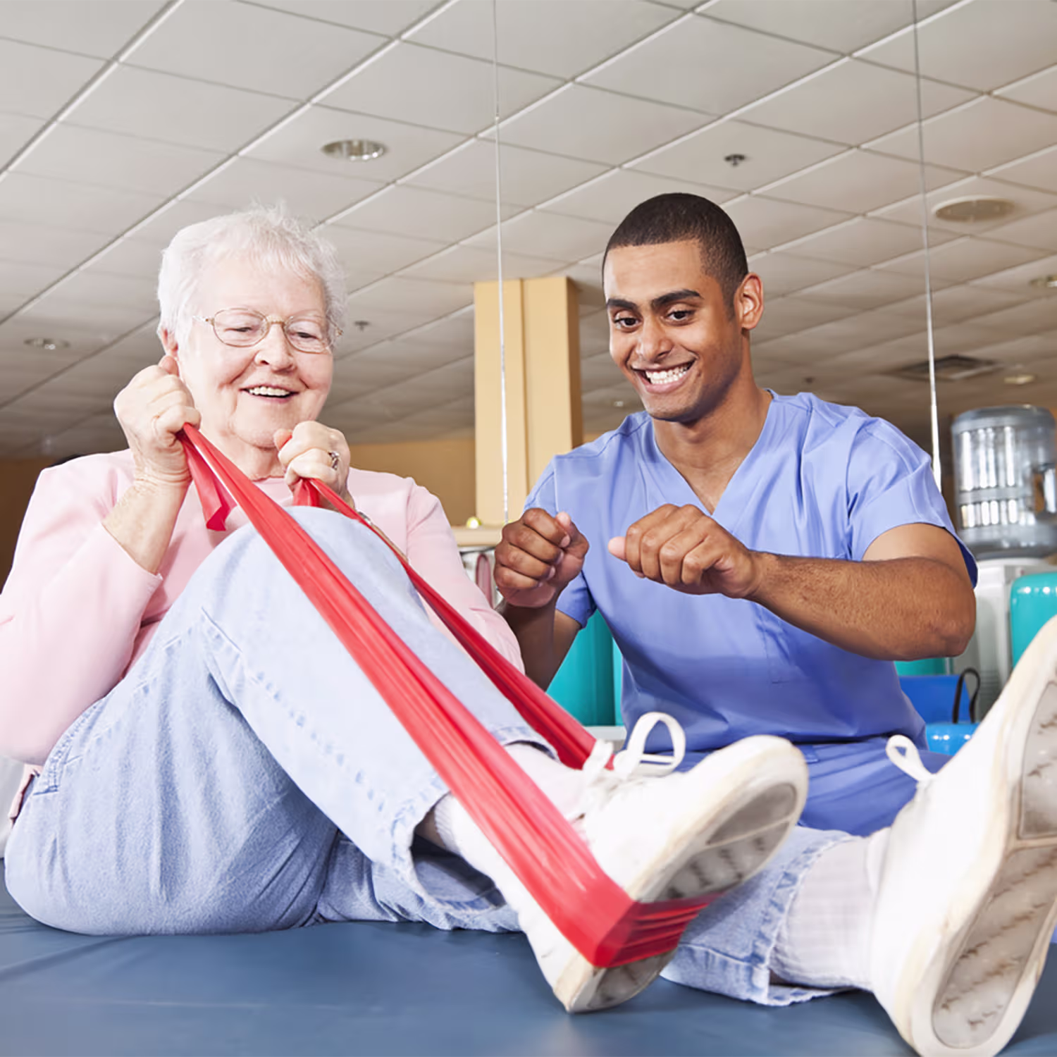 Senior woman smiling and exercising her legs with a red resistance band assisted by a male physical therapist in blue scrubs.