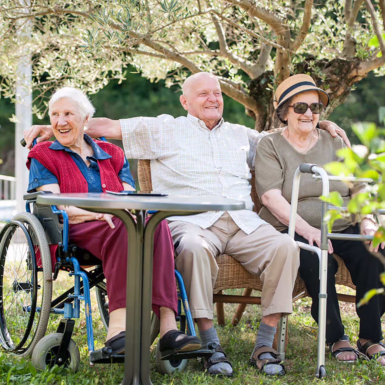 Three elderly people smiling outdoors, one woman in a wheelchair, a man sitting on a wicker chair with arms around both women, and another woman with sunglasses and a hat using a walker.
