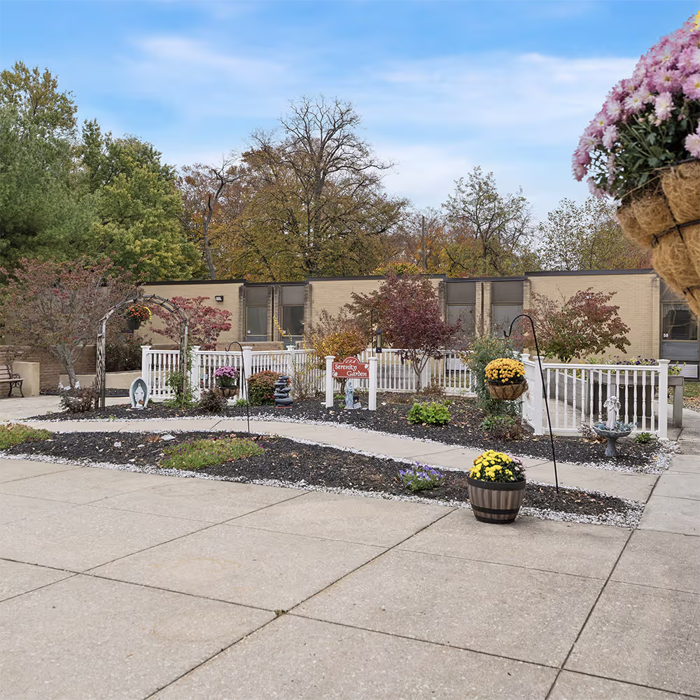 Outdoor garden area with pathway, white railing, colorful flowers in pots, and trees against a blue sky.