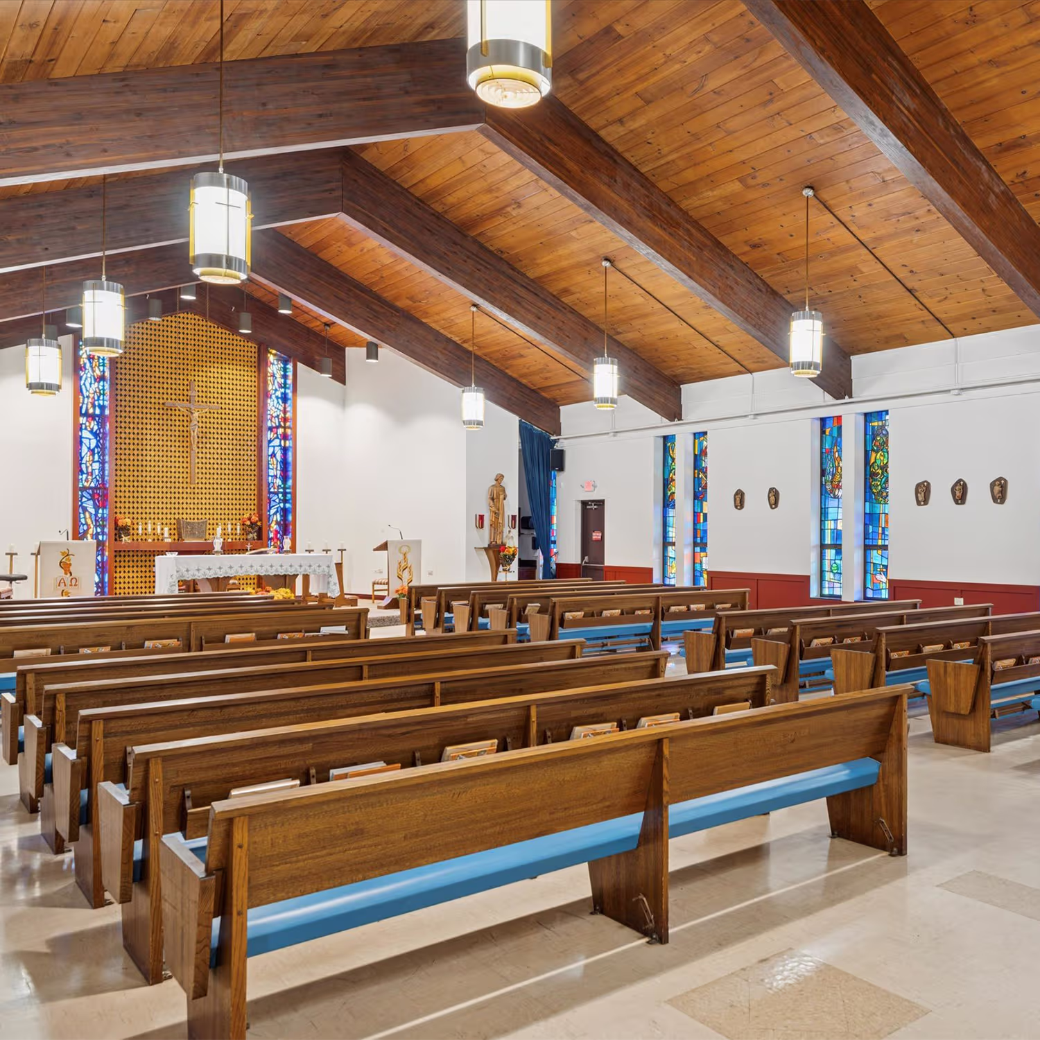 Interior of a church with wooden pews, a wooden ceiling with exposed beams, hanging pendant lights, stained glass windows, and an altar with a crucifix and statues.